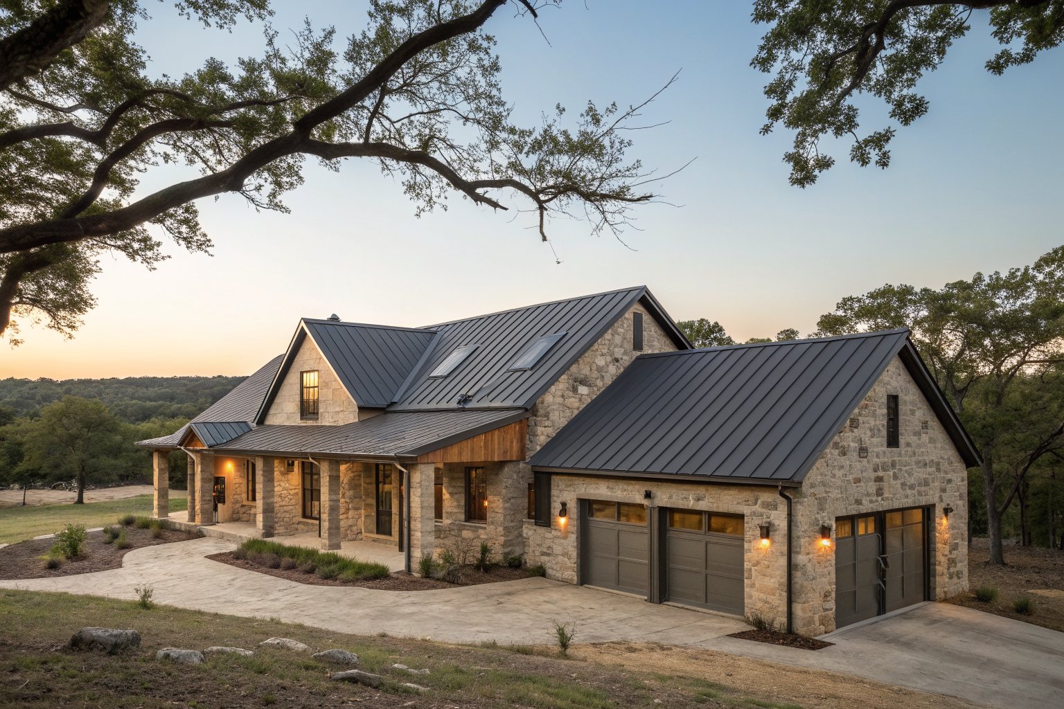 A large stone house with a dark standing-seam metal roof and a three-car garage among oaks at dusk
