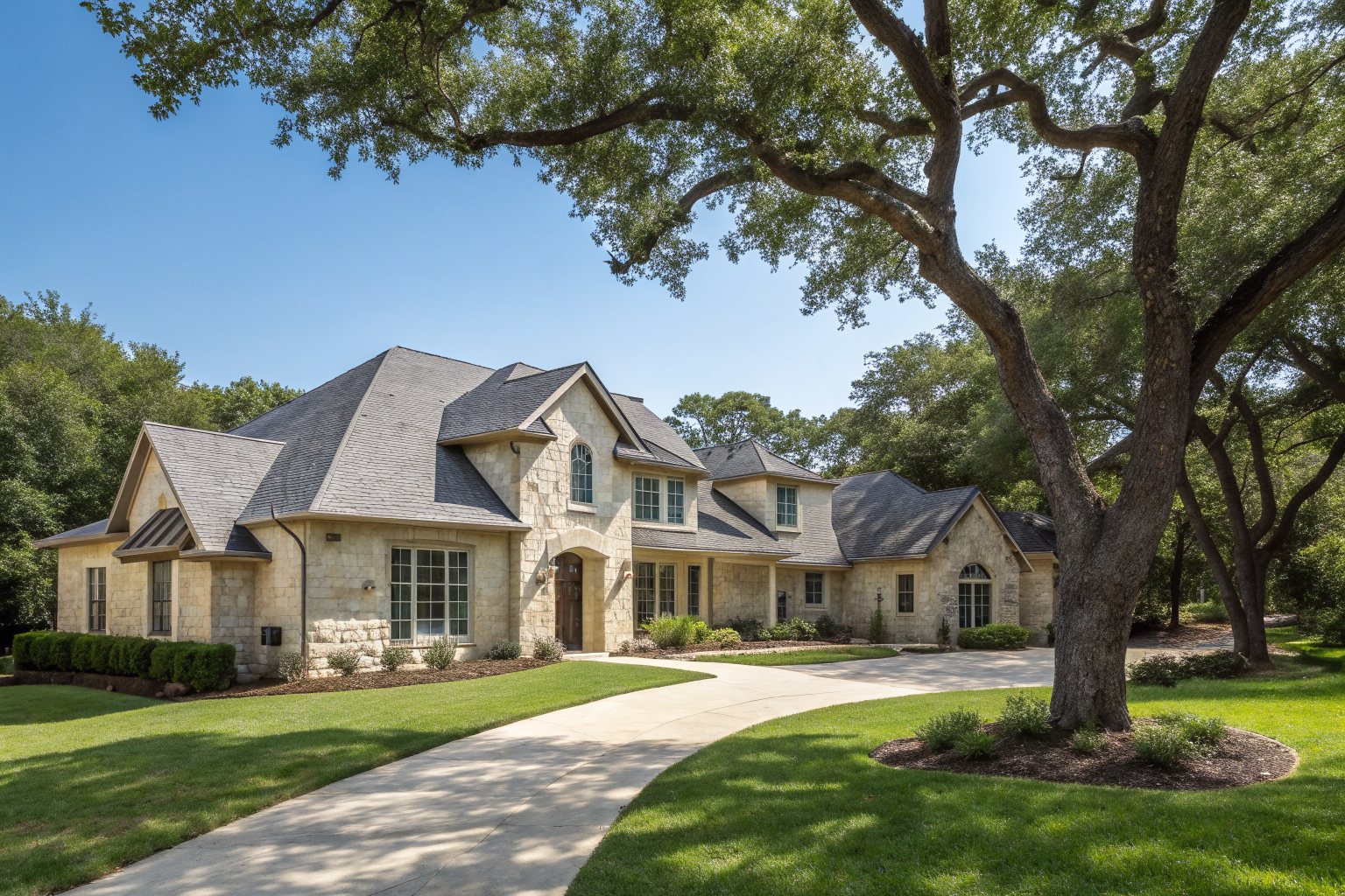 A two-story cream limestone house with a charcoal shingle roof under a large live oak, with a curving driveway