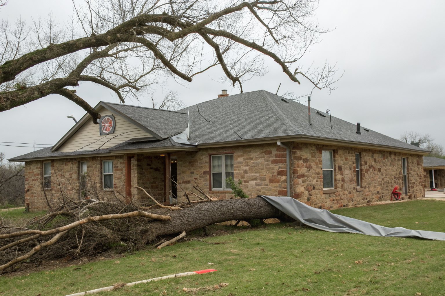 A fallen oak limb across the lawn of a stone ranch house after a storm, with a gray tarp spread on the grass